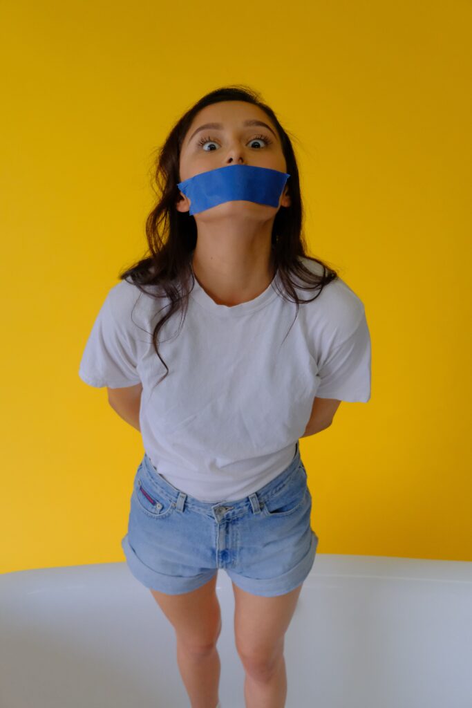 Image of a girl with her hands behind her back, tape over her month, with the expression that she's trying to say something. Yellow background.
