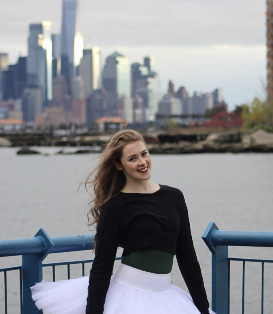 Photo of Ellington smiling while wearing a white tutu, forest green leotard, and black long sleeve jacket with the NYC skyline in the background.